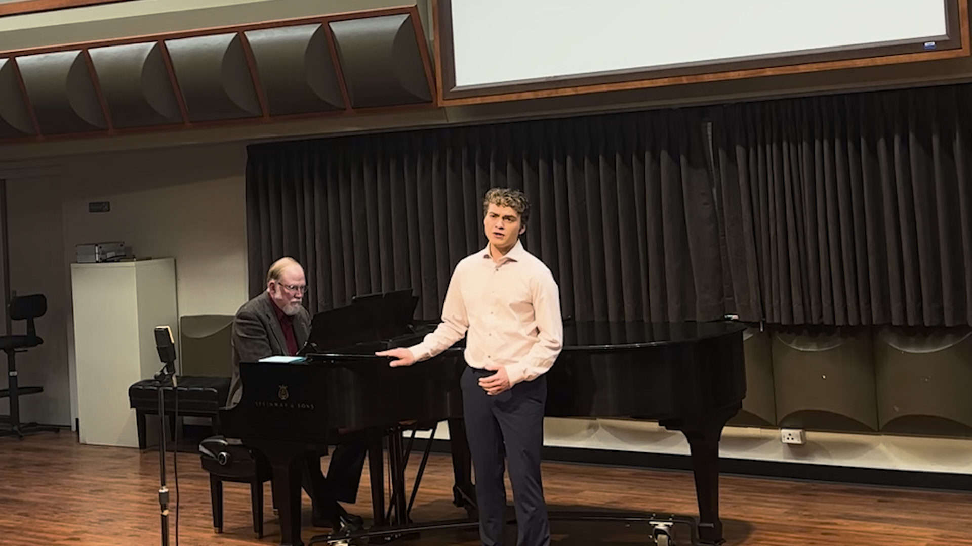 Soloist wearing pink dress shirt black pants, infront of black piano with pianist dressed in all black